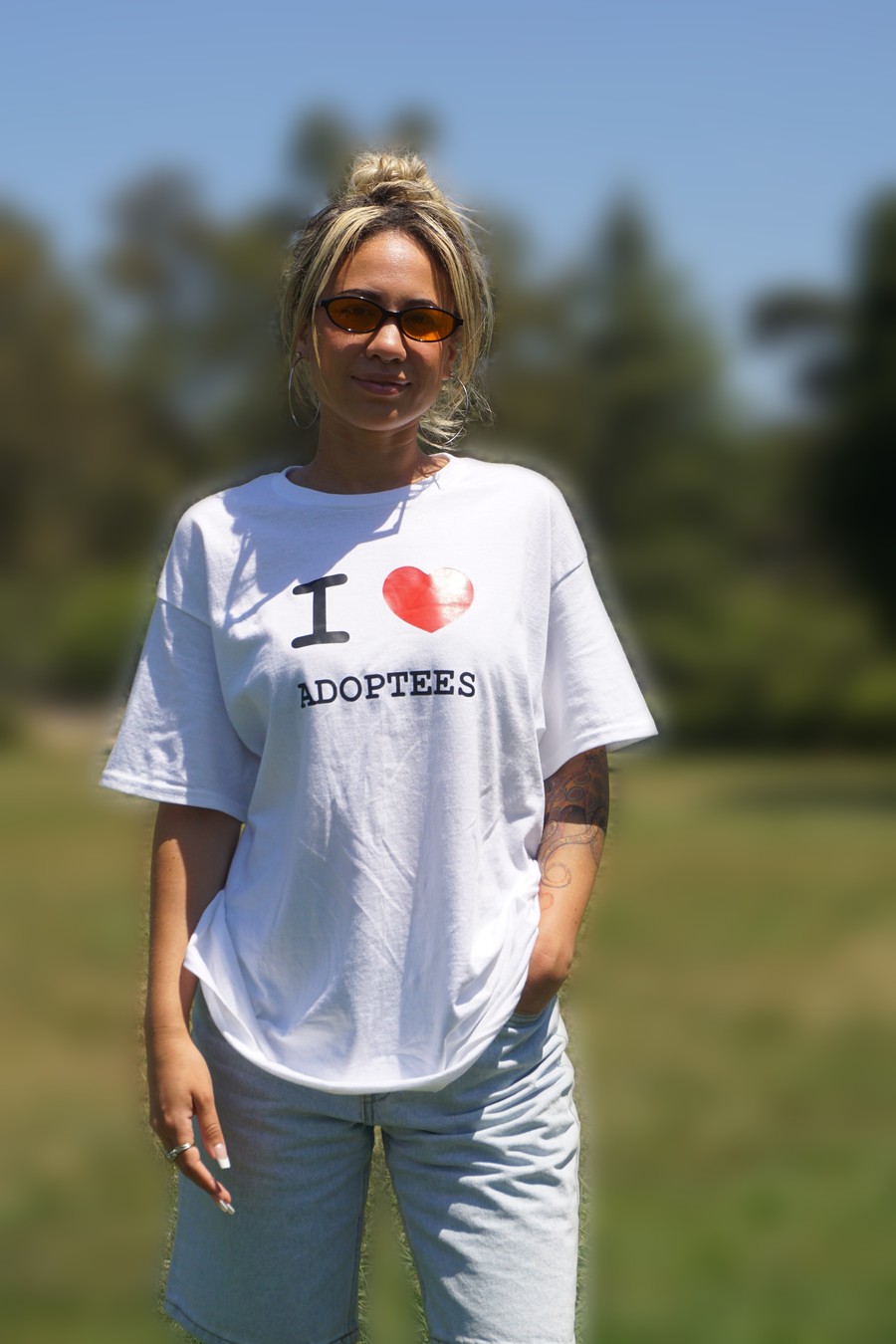 Alexis Kimball standing with one hand in her pocket, wearing sunglasses and a white t shirt reading I heart adoptees.